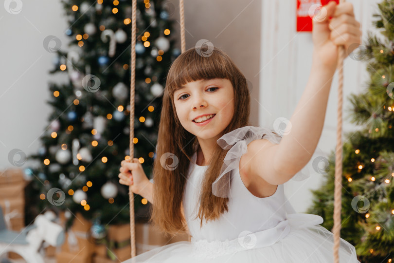 Скачать счастливая маленькая девочка в красивом белом платье сидит на качелях возле рождественской елки в студии. новый год. фотосток Ozero