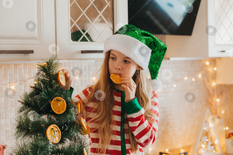Скачать Маленькая девочка в шапочке Санта-Клауса украшает елку половинками сушеного апельсина. Милый малыш готовится отпраздновать Рождество. фотосток Ozero