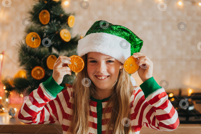 Скачать Маленькая улыбающаяся симпатичная блондинка в пижаме и оленьих рогах подносит половинки фруктов к глазам. ребенок на кухне возле рождественской елки. Рождество фотосток Ozero