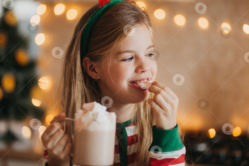 Скачать маленькая белокурая девочка в полосатой пижаме держит кружку с горячим напитком и зефиром на украшенной кухне. зимние выходные и праздничные дни. место для текста. фотосток Ozero