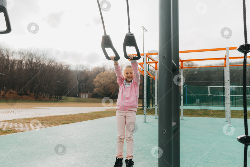 Скачать Маленькая веселая девочка играет на детской площадке. Ребенок занимается на гимнастических кольцах на улице. Ребенок занимается спортом на тренажерах. фотосток Ozero