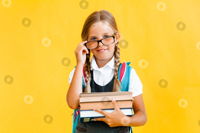Скачать Портрет милой маленькой девочки на желтом фоне. Школьница смотрит в камеру, держа в руках книгу и поправляя очки. Концепция образования. фотосток Ozero
