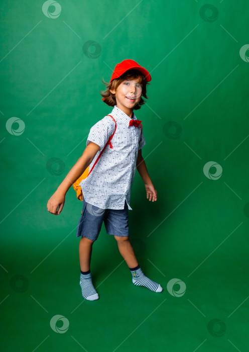 Скачать Маленький эмоциональный 7-летний мальчик в красной шапочке и с желтым рюкзачком. Стою на зеленом фоне, развлекаюсь. фотосток Ozero