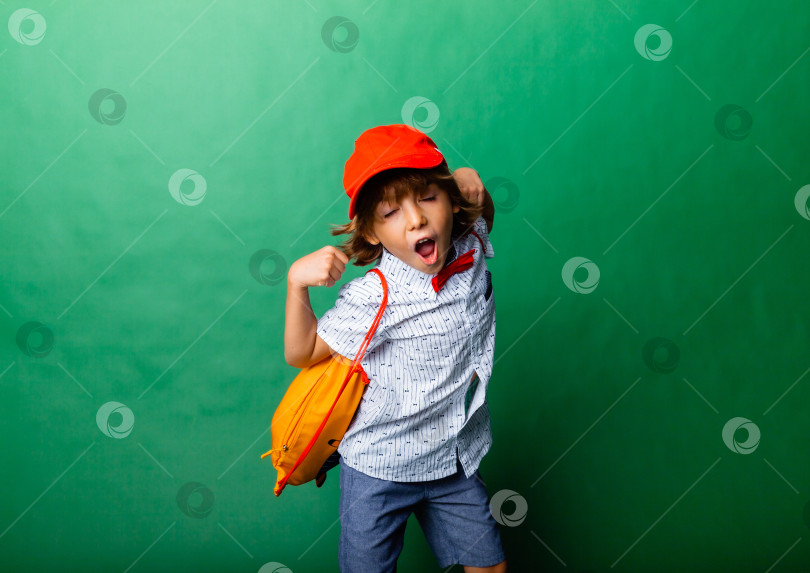 Скачать Маленький эмоциональный 7-летний мальчик в красной шапочке и с желтым рюкзачком. Стою на зеленом фоне, развлекаюсь. фотосток Ozero