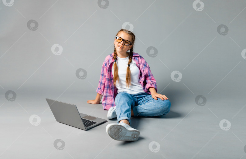 Скачать портрет девочки-школьницы, использующей ноутбук, сидя со скрещенными ногами, изолированный на сером фоне фотосток Ozero