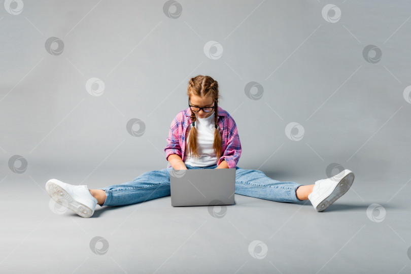 Скачать портрет девочки-школьницы, использующей ноутбук, сидя со скрещенными ногами, изолированный на сером фоне фотосток Ozero