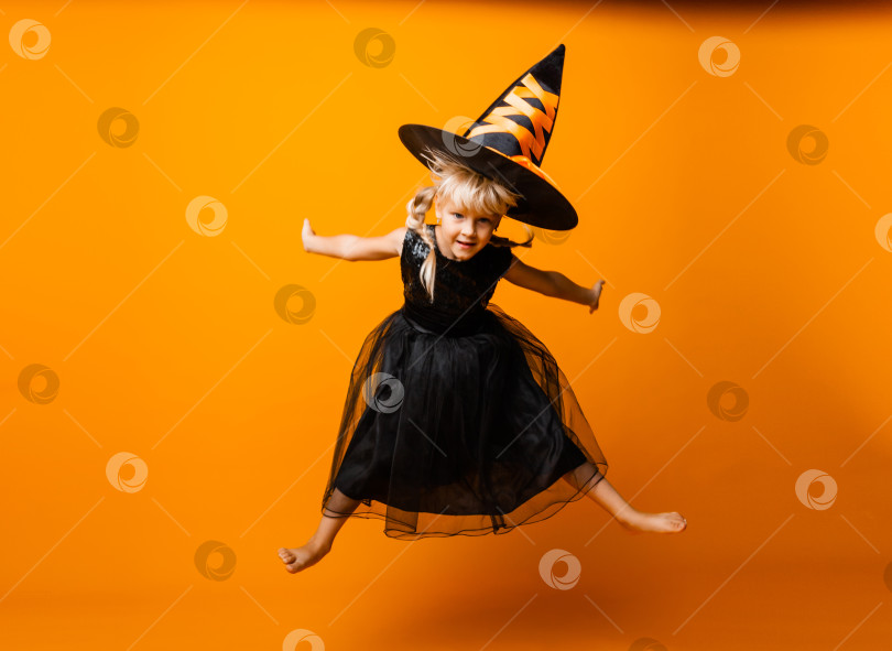 Скачать Маленькая девочка в черном костюме на Хэллоуин смеется и смотрит в камеру, прыгает и веселится, изолированная на желтом фоне. Хэллоуин фотосток Ozero