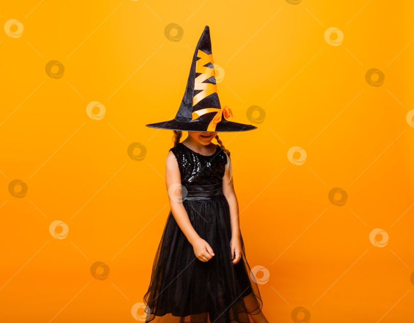 Скачать Маленькая девочка в черном костюме на Хэллоуин смеется и смотрит в камеру, прыгает и веселится, изолированная на желтом фоне. Хэллоуин фотосток Ozero