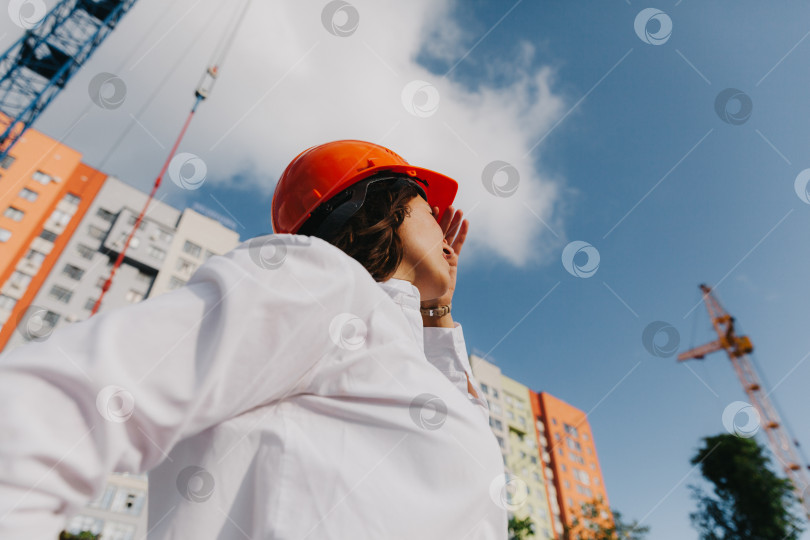 Скачать Молодая красивая женщина-архитектор в белой рубашке и каске держит в руках доску для переворачивания. женщина в каске на строительной площадке у дома. фотосток Ozero