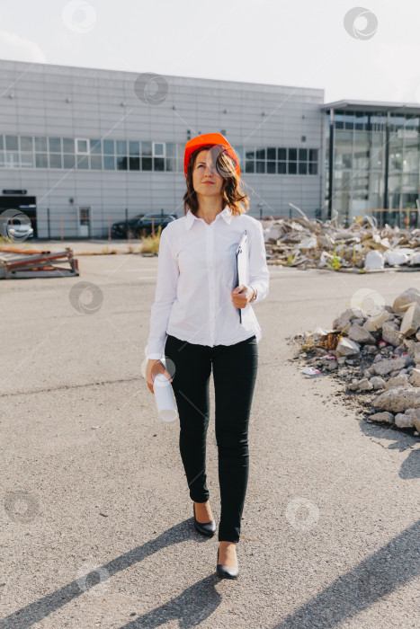 Скачать Молодая красивая женщина-архитектор в белой рубашке и каске держит в руках доску для переворачивания. женщина в каске на строительной площадке у дома. фотосток Ozero