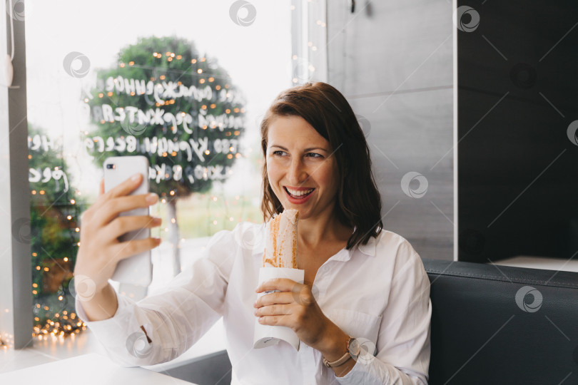 Скачать Счастливая девушка делает селфи на мобильный телефон, поедая твистер в кафе. фотосток Ozero