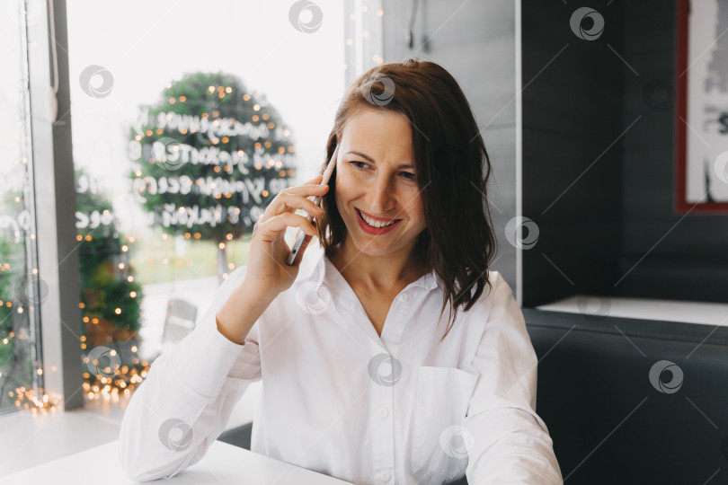 Скачать Молодая счастливая женщина разговаривает по мобильному телефону в кафе, дама с красивой улыбкой разговаривает по мобильному телефону, сидя в кафе во время обеда фотосток Ozero