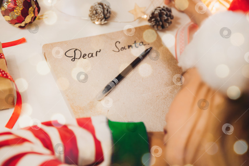 Скачать Маленькая девочка перед Рождеством пишет письмо Деду Морозу на столе с гирляндами. ребенок пишет письмо Деду Морозу. фотосток Ozero