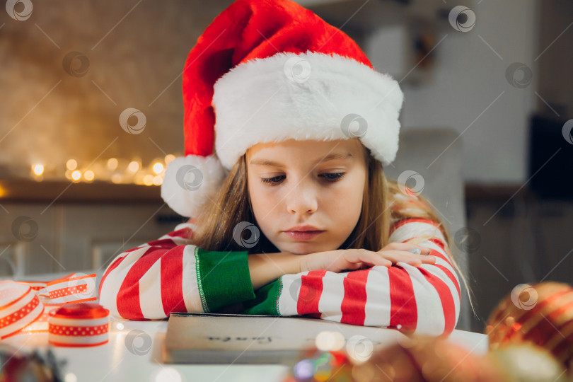Скачать Маленькая девочка перед Рождеством пишет письмо Деду Морозу на столе с гирляндами. ребенок пишет письмо Деду Морозу. фотосток Ozero