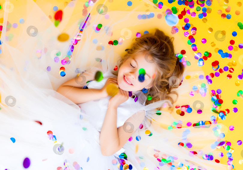 Скачать Счастливая маленькая девочка в белом платье празднует вечеринку с конфетти, вид сверху. День рождения ребенка, положительные эмоции. фотосток Ozero