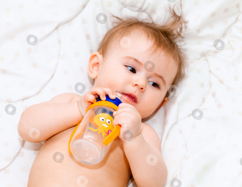 Скачать ребенок в возрасте 6 месяцев пьет воду из бутылочки, лежа на кровати. счастливый и радостный малыш фотосток Ozero