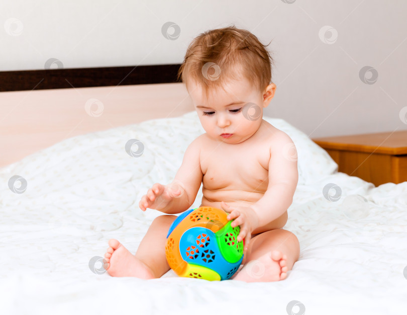 Скачать Симпатичная белокурая малышка играет с мячом, сидя на кровати в спальне. счастливый малыш 6 месяцев от роду играет с мячом фотосток Ozero