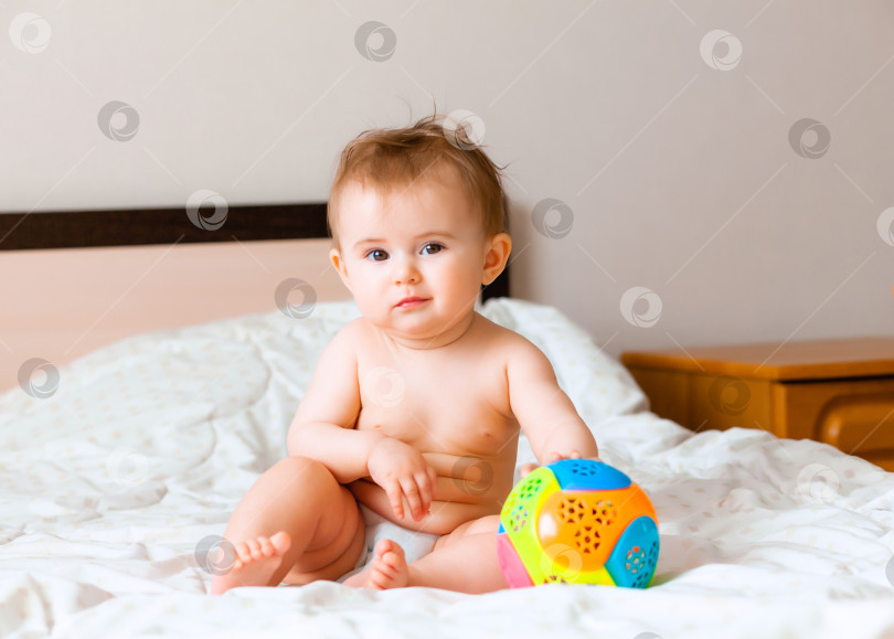 Скачать Симпатичная белокурая малышка играет с мячом, сидя на кровати в спальне. счастливый малыш 6 месяцев от роду играет с мячом фотосток Ozero