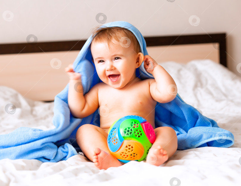 Скачать Симпатичный белокурый малыш, завернутый в голубое банное полотенце, играет с мячом, сидя на кровати в спальне. счастливый 6-месячный малыш играет с мячом фотосток Ozero