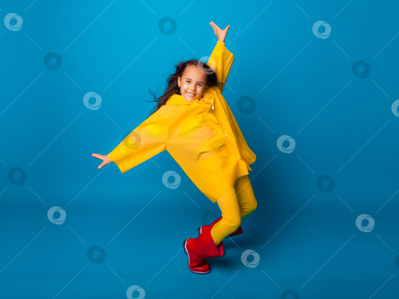 Скачать Веселая девушка в желтом плаще и красных резиновых сапогах прыгает с раскинутыми руками и смотрит в камеру. фотосток Ozero