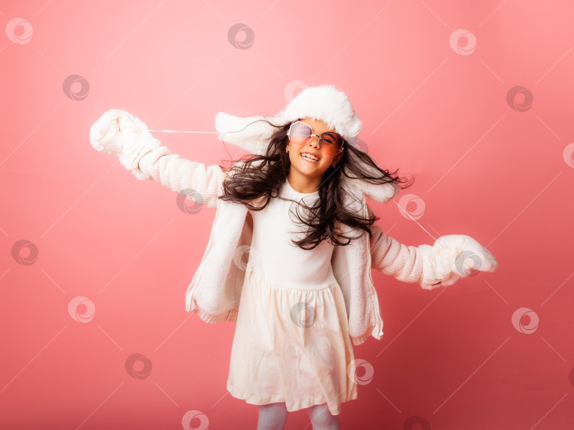 Скачать Маленькая девочка-брюнетка с длинными волосами в зимней меховой шапке и варежках на розовом фоне. фотосток Ozero