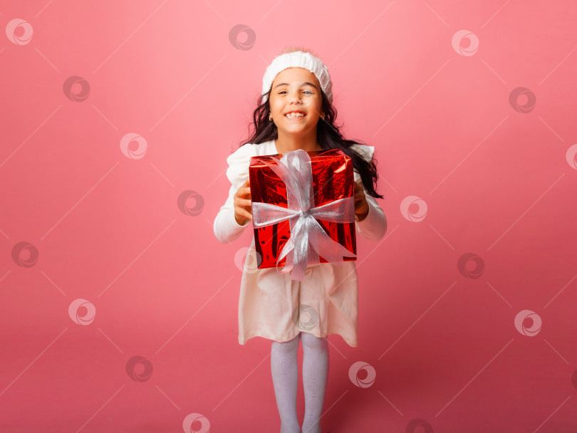 Скачать маленькая брюнетка с длинными волосами в зимней шапочке держит подарок на розовом фоне. фотосток Ozero