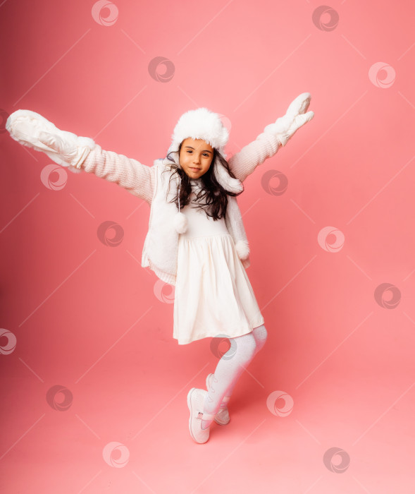 Скачать Маленькая девочка-брюнетка с длинными волосами в зимней меховой шапке и варежках на розовом фоне. фотосток Ozero
