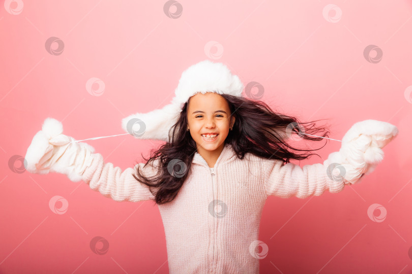 Скачать Маленькая девочка-брюнетка с длинными волосами в зимней шапочке и варежках на розовом фоне фотосток Ozero