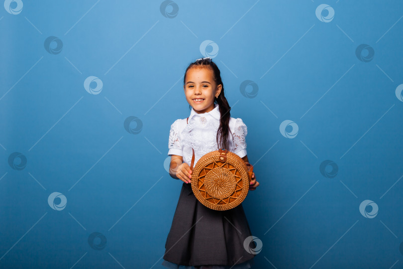 Скачать Симпатичная маленькая девочка семи лет позирует в школьной форме и держит в руках плетеную круглую сумку на синем фоне фотосток Ozero