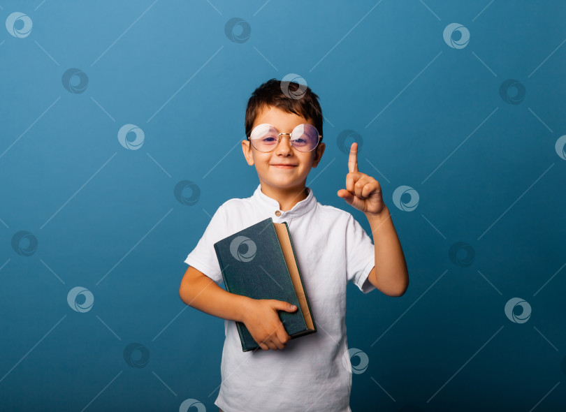 Скачать Веселый мальчик кавказской внешности держит в руках книгу. мальчик с книгой указывает пальцем вверх на синем фоне. фотосток Ozero