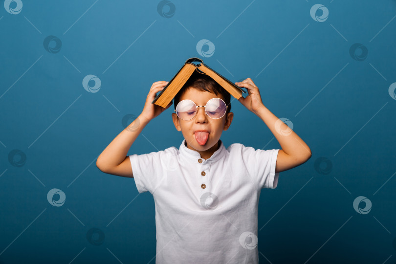 Скачать Симпатичный мальчик в очках держит книгу на голове, на синем фоне. мальчик с книгой показывает язык. школьник. фотосток Ozero