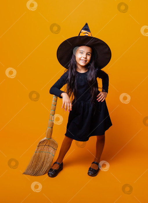 Скачать Маленькая девочка, одетая как ведьма на Хэллоуин, в черном платье и шляпе, летит на метле на желтом фоне. фотосток Ozero