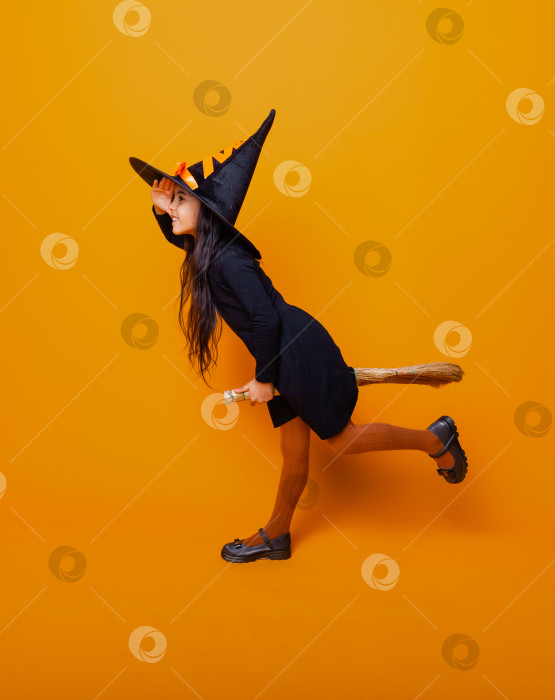 Скачать Маленькая девочка, одетая как ведьма на Хэллоуин, в черном платье и шляпе летает на метле на желтом фоне. фотосток Ozero