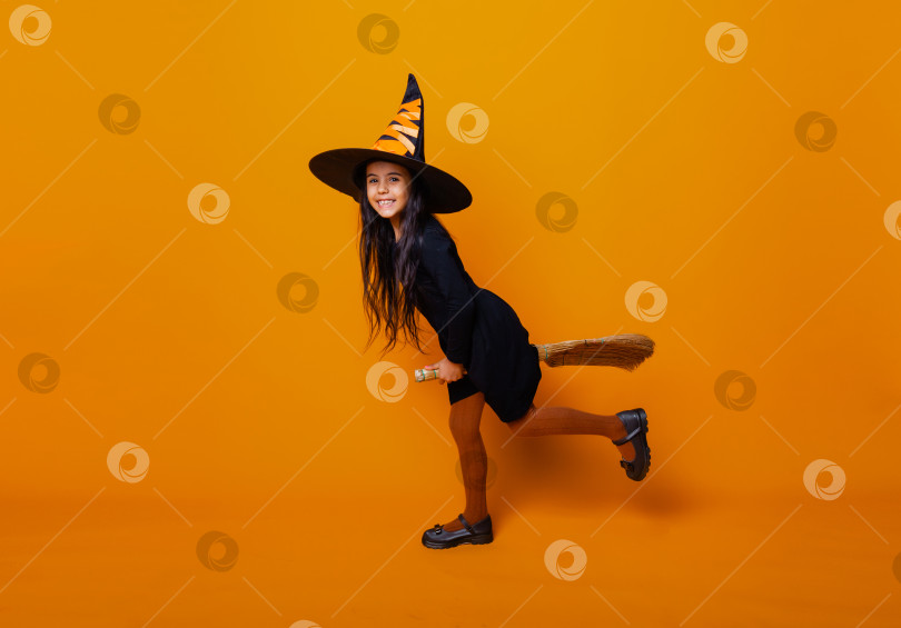 Скачать Маленькая девочка, одетая как ведьма на Хэллоуин, в черном платье и шляпе, летит на метле на желтом фоне. фотосток Ozero
