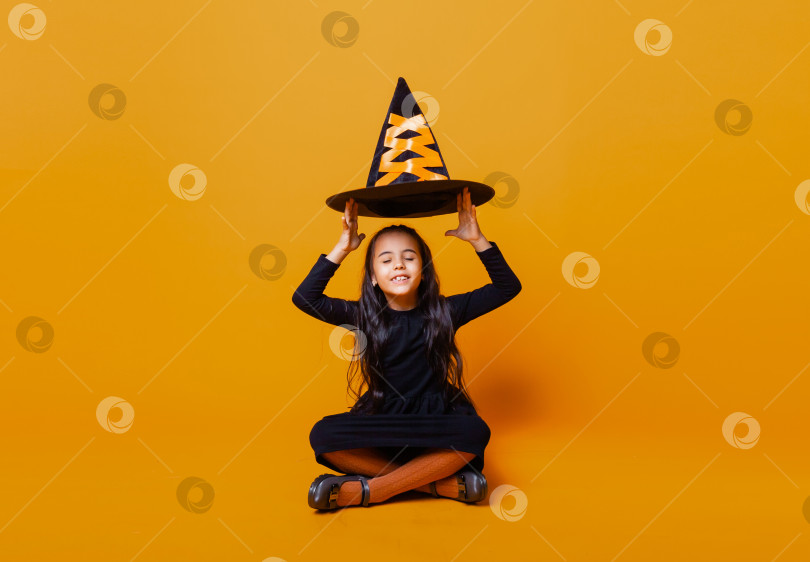 Скачать Хэллоуин. веселая маленькая ведьма сидит на желтом фоне и держит в руках шляпу ведьмы на Хэллоуин. фотосток Ozero
