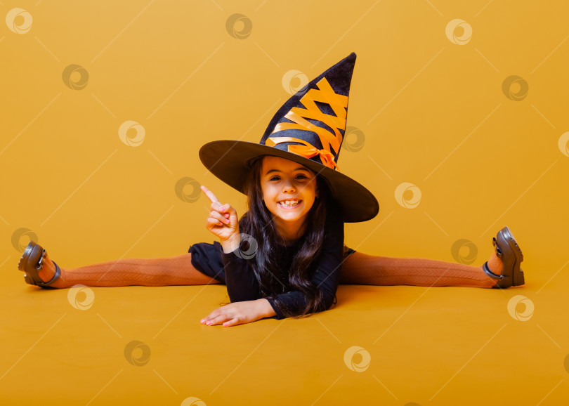 Скачать маленькая девочка в костюме ведьмы и в шляпе сидит на шпагате на желтом фоне с пробелом для копирования. хэллоуин. фотосток Ozero