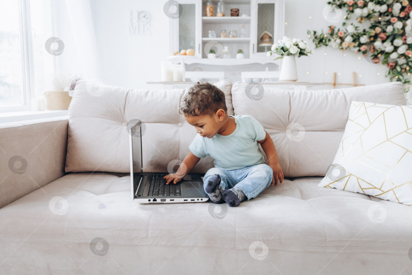 Скачать маленький темнокожий мальчик-афроамериканец сидит на диване с ноутбуком. фотосток Ozero