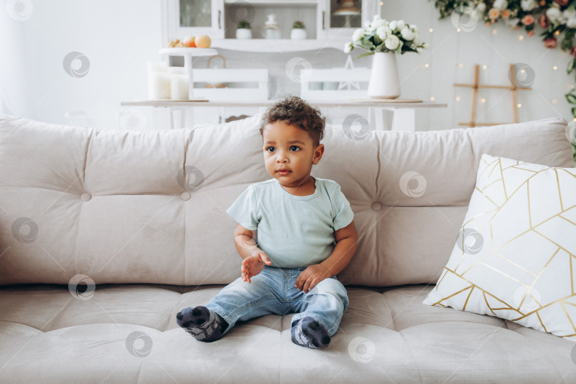 Скачать Симпатичный чернокожий мальчик-афроамериканец сидит на диване в гостиной. фотосток Ozero