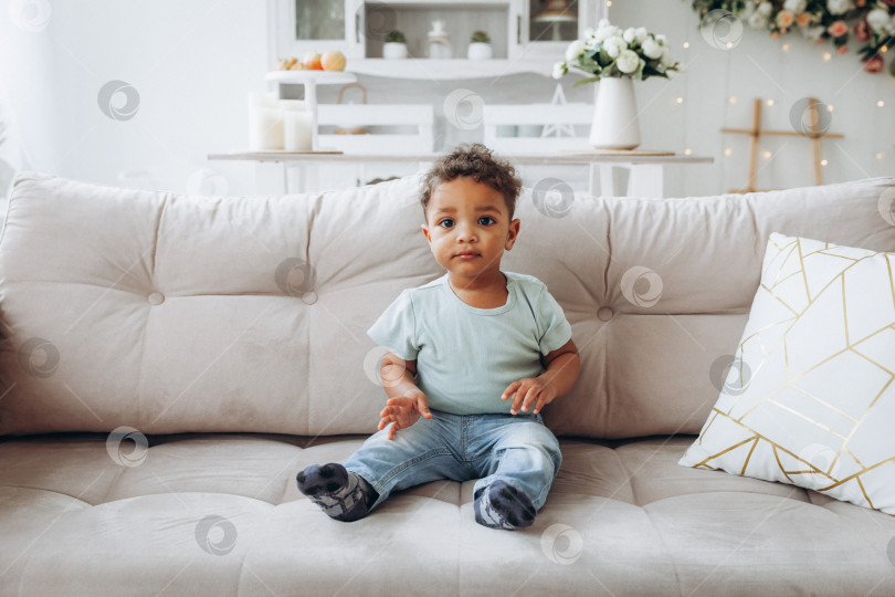 Скачать Симпатичный чернокожий мальчик-афроамериканец сидит на диване в гостиной. фотосток Ozero