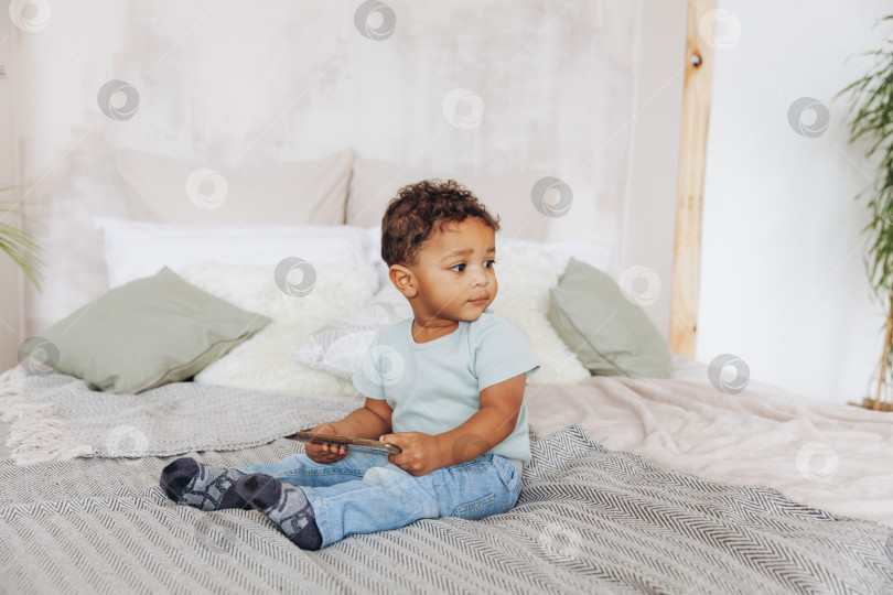 Скачать Милый маленький афроамериканский мальчик со смартфоном дома на кровати, счастливый ребенок смотрит мультфильмы по телефону. фотосток Ozero