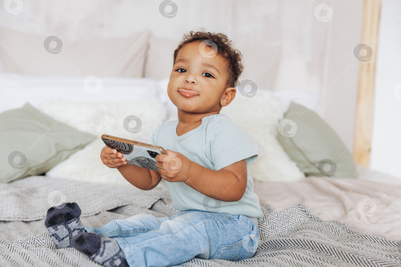 Скачать Милый маленький афроамериканский мальчик со смартфоном дома на кровати, счастливый ребенок смотрит мультфильмы по телефону. фотосток Ozero