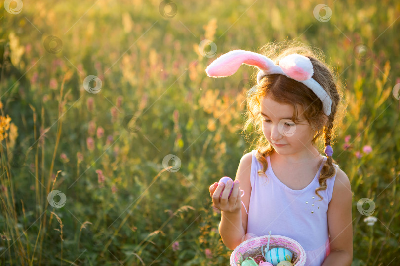 Скачать Милая забавная девочка с расписными пасхальными яйцами весной на природе в поле с золотым солнечным светом и цветами. Пасхальный праздник, пасхальный кролик с ушами, разноцветные яйца в корзинке. Образ жизни фотосток Ozero