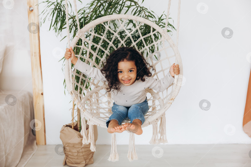 Скачать маленькая афроамериканская девочка в плетеном кресле, висящем в гостиной. фотосток Ozero