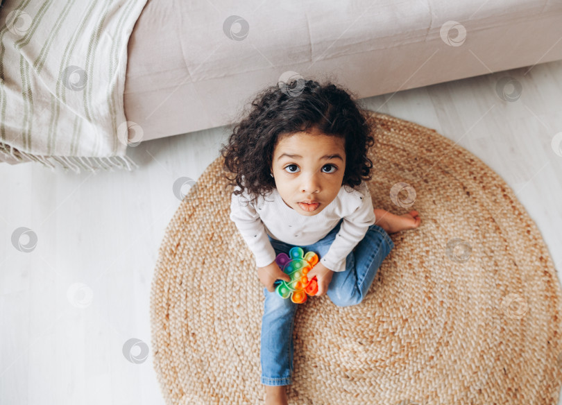 Скачать Афроамериканская девочка дома на полу играет с поп-игрушкой, нажимает на кнопки, снимает стресс фотосток Ozero