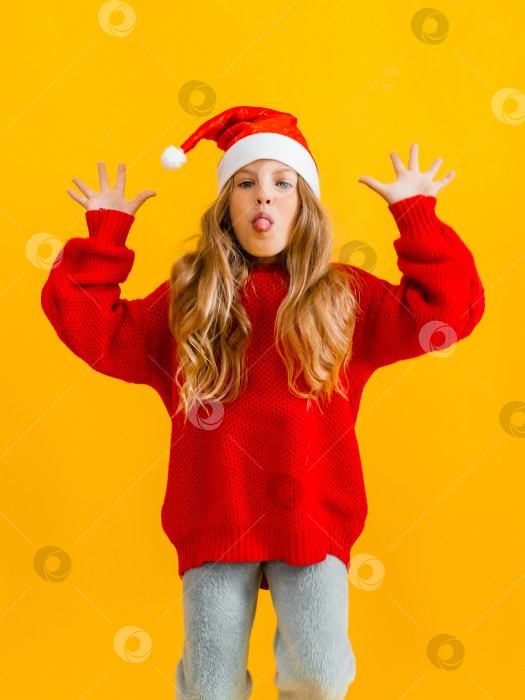 Скачать маленькая девочка в шапочке Санты и красном свитере прыгает и показывает пятерку на желтом фоне. фотосток Ozero
