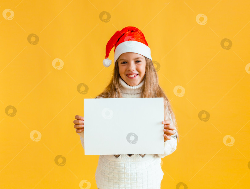 Скачать Маленькая девочка 8 лет со светлыми волосами в шапочке Санта-Клауса держит белый лист бумаги на желтом фоне. фотосток Ozero