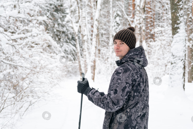 Скачать Лыжник в шапке с помпоном, с лыжными палками в руках, спиной на фоне заснеженного леса. Катание на беговых лыжах в зимнем лесу, спорт на открытом воздухе, здоровый образ жизни. фотосток Ozero