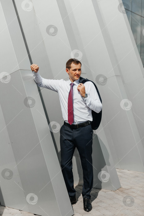 Скачать молодой мужчина-бизнесмен, идущий домой с работы с курткой на плече, улыбается и позирует возле офисного здания фотосток Ozero