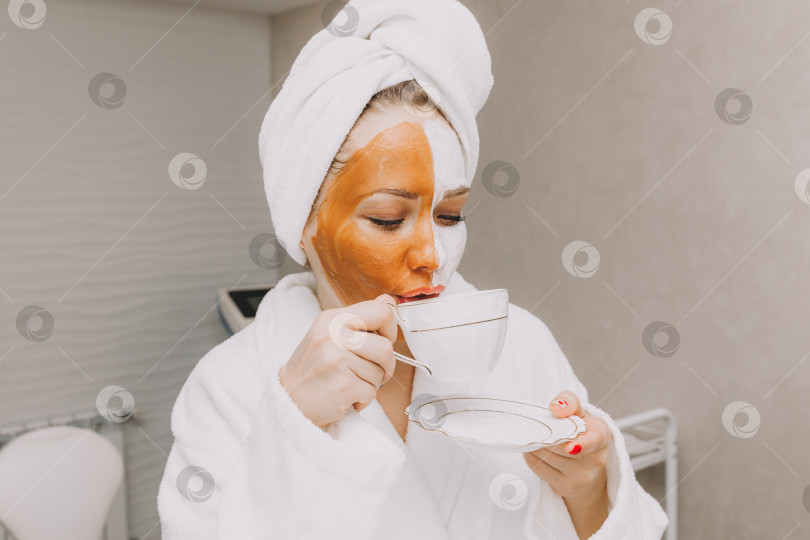 Скачать Красивая молодая женщина с маской на лице пьет кофе в кабинете косметолога. фотосток Ozero
