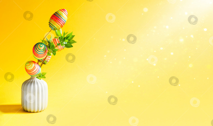 Скачать Букет из зеленых веток с распускающимися свежими листьями в вазе украшен разноцветными радужными пасхальными яйцами. Декор в интерьере дома к Пасхе. Пространство для копирования. Желтый пасхальный фон фотосток Ozero